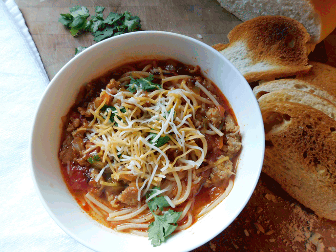 White bowl with sausage and noodles in broth on a chopping block with toasted bread