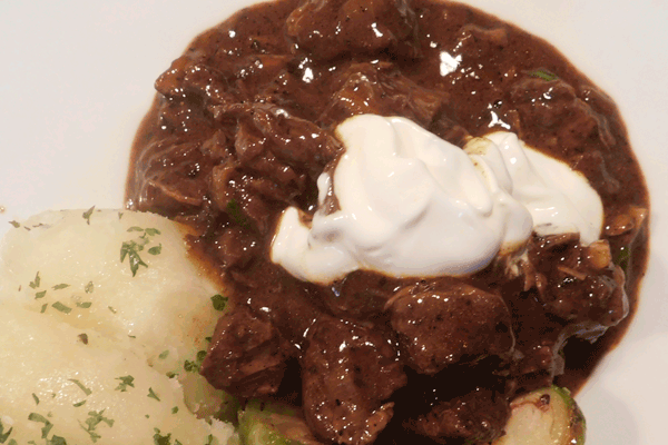 Beef stew on white plate with sour cream on top and herbed potatoes next to it