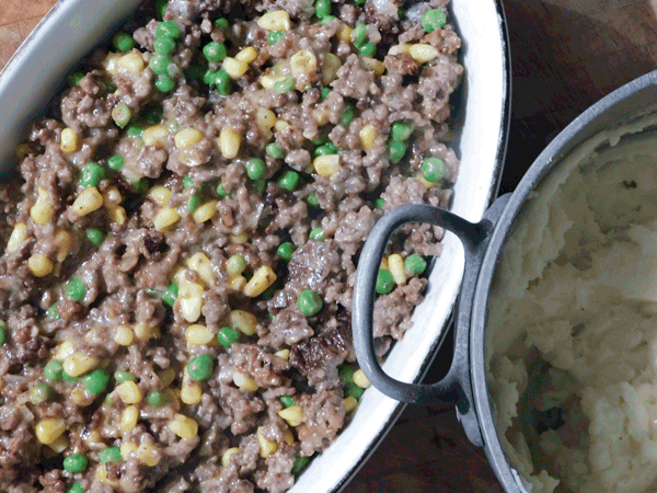 Cooked ground beef and veggies in a casserole dish with a pot of mashed potatoes next to it