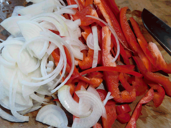 sliced red peppers and onions on a chopping block with a knife next to them
