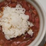 Red beans with a scoop of white rice in a white bowl next to a piece of cooked sausage