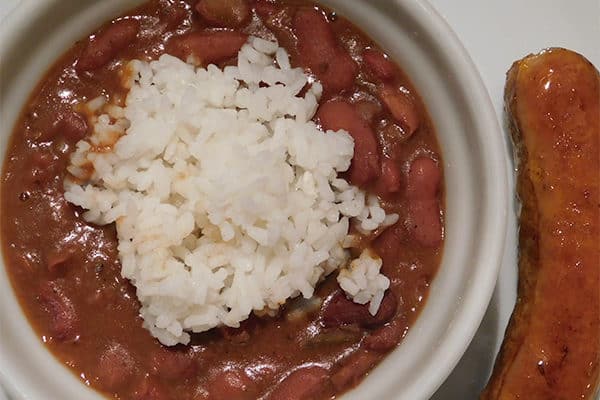 Red beans with a scoop of white rice in a white bowl next to a piece of cooked sausage
