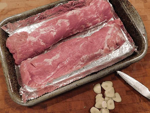 Two Pork tenderloins on a baking dish with some garlic sliced next to it on a chopping board