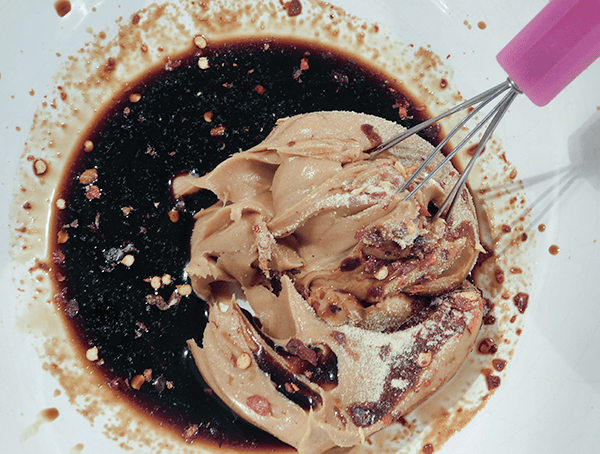 Peanut butter, soy sauce red pepper flakes and garlic powder in white bowl with a pink whisk