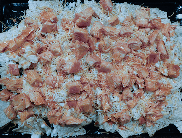 Buffalo CHicken Nachos = Corn chips on a sheet pan with cheese and sauced chicken ready for the oven
