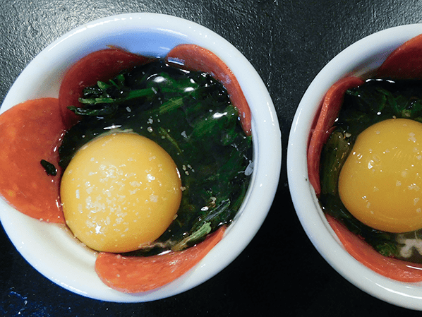 Closeup of two white ramekins with pepperoni, spinach and uncooked eggs