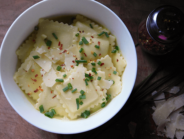 Ravioli in Broth in a white bowl next to pepper flakes, chives and parmesan cheese
