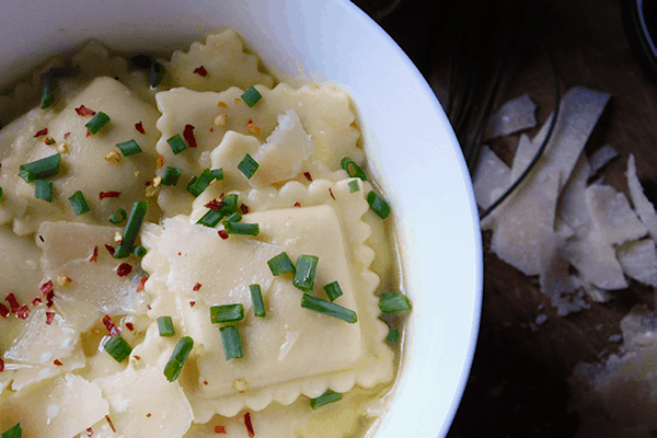 Ravioli in Broth in white bowl with parmesan cheese in background